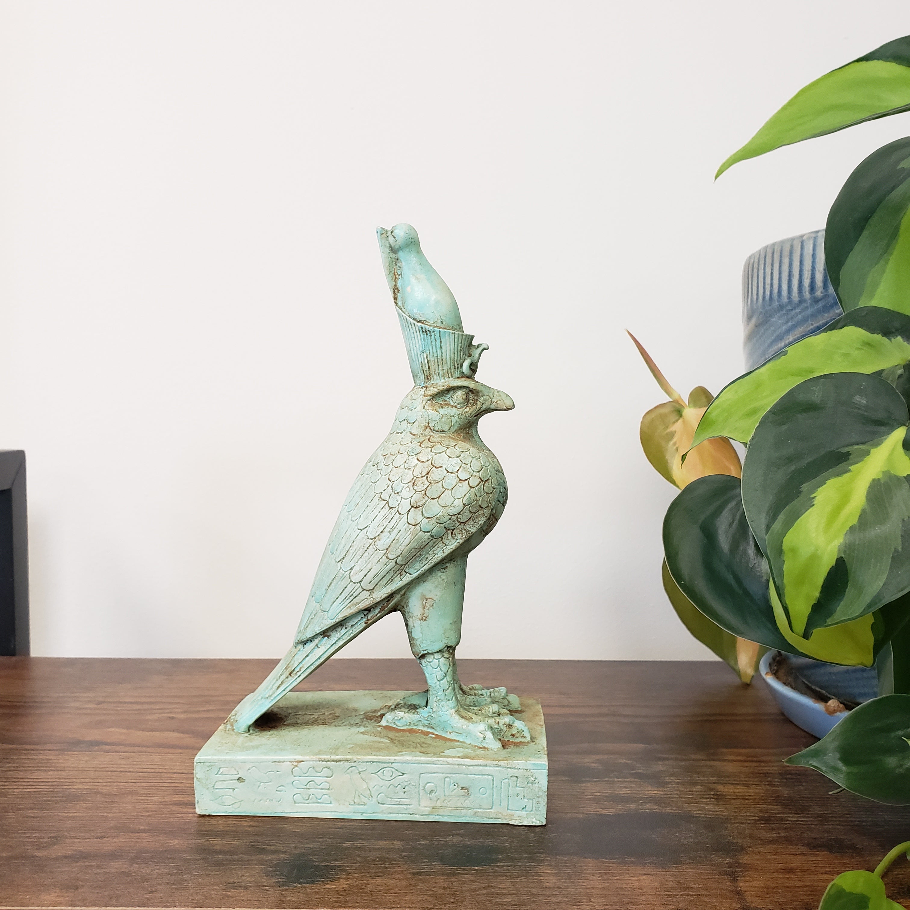 Decorative green Egyptian falcon-god Horus sculpture on a wooden surface with a plant in the background