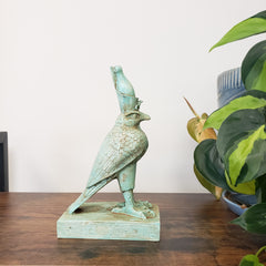 Decorative green Egyptian falcon-god Horus sculpture on a wooden surface with a plant in the background