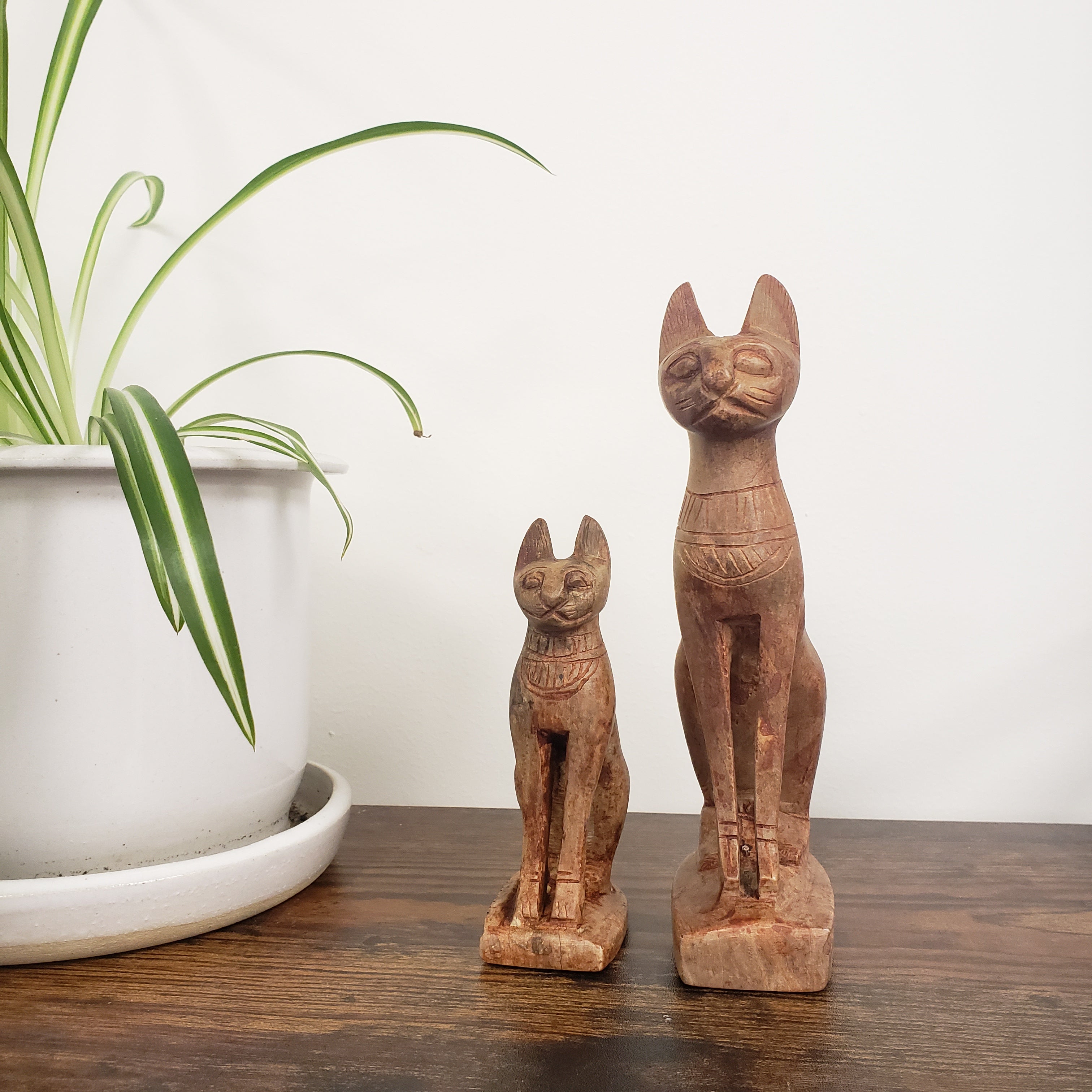Two stone cat figurines on a shelf with a plant and white container in the background.
