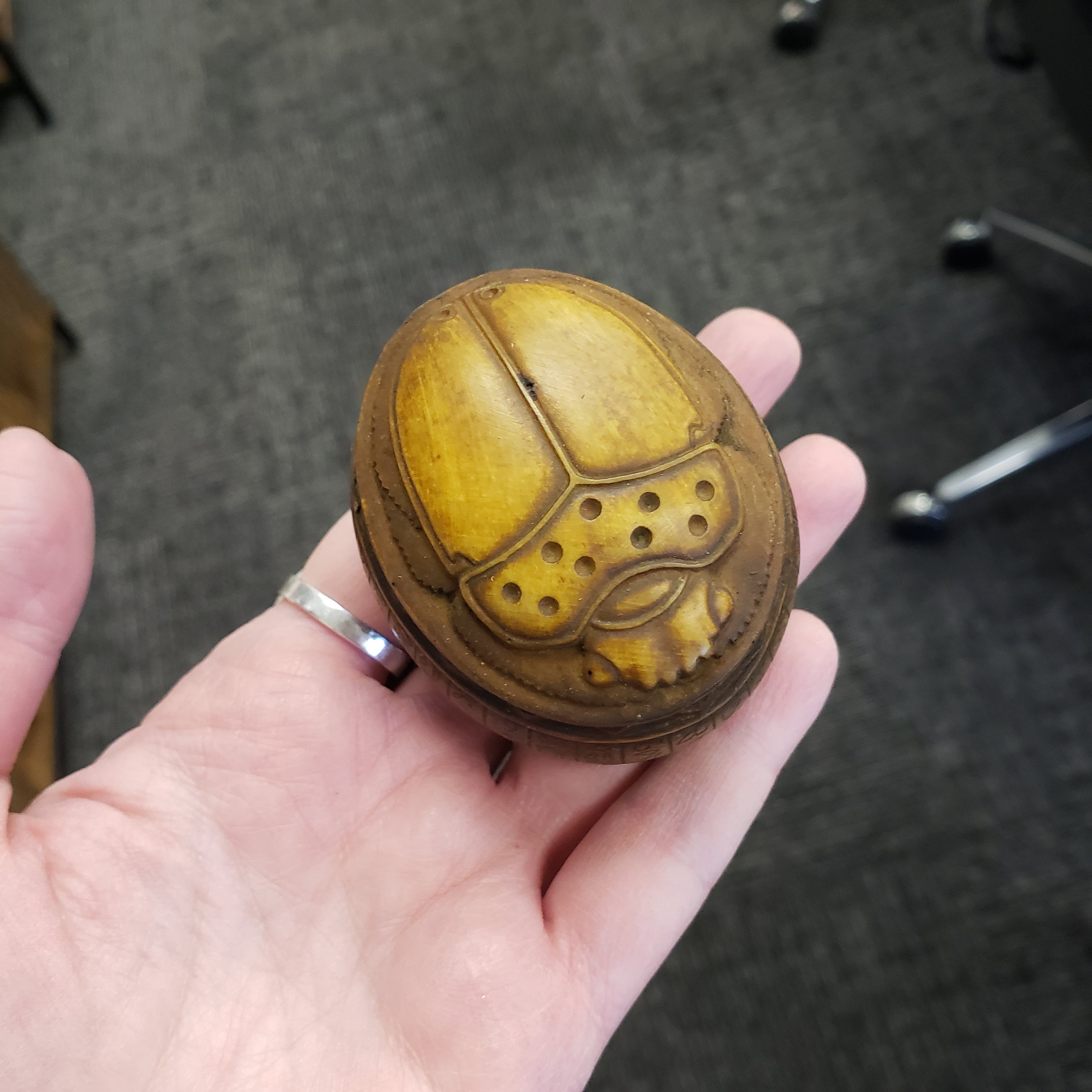 Hand holding a round, brown object with a detailed design on a gray carpeted floor.