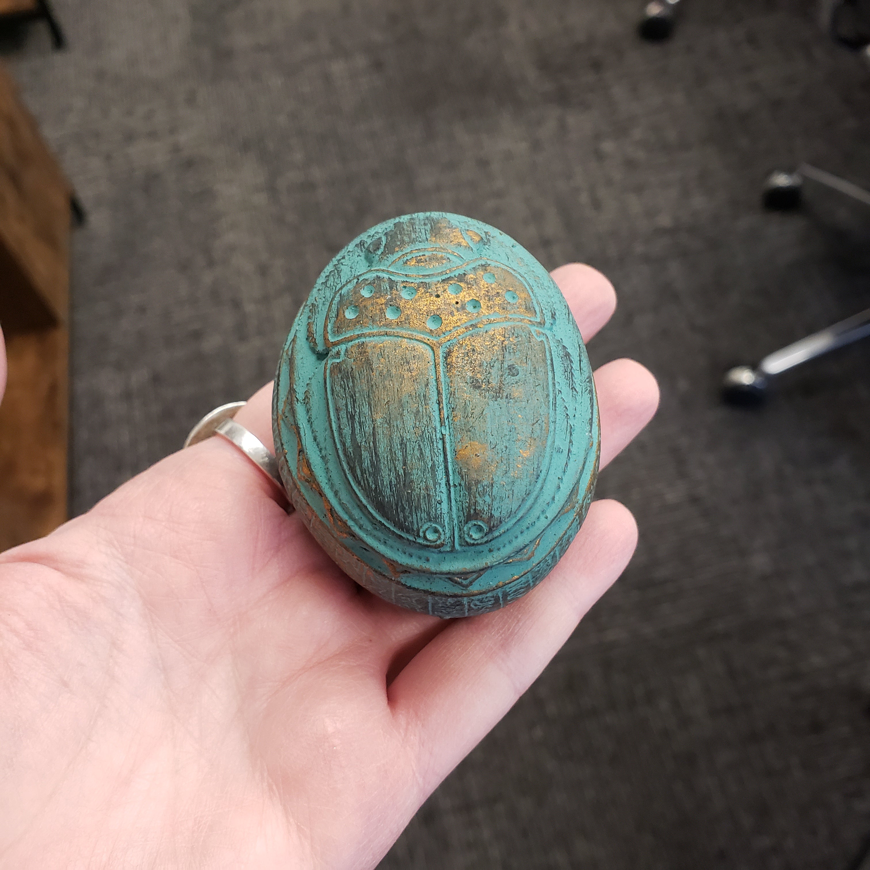 Hand holding a greenish-blue decorative object with intricate patterns on a carpeted floor.