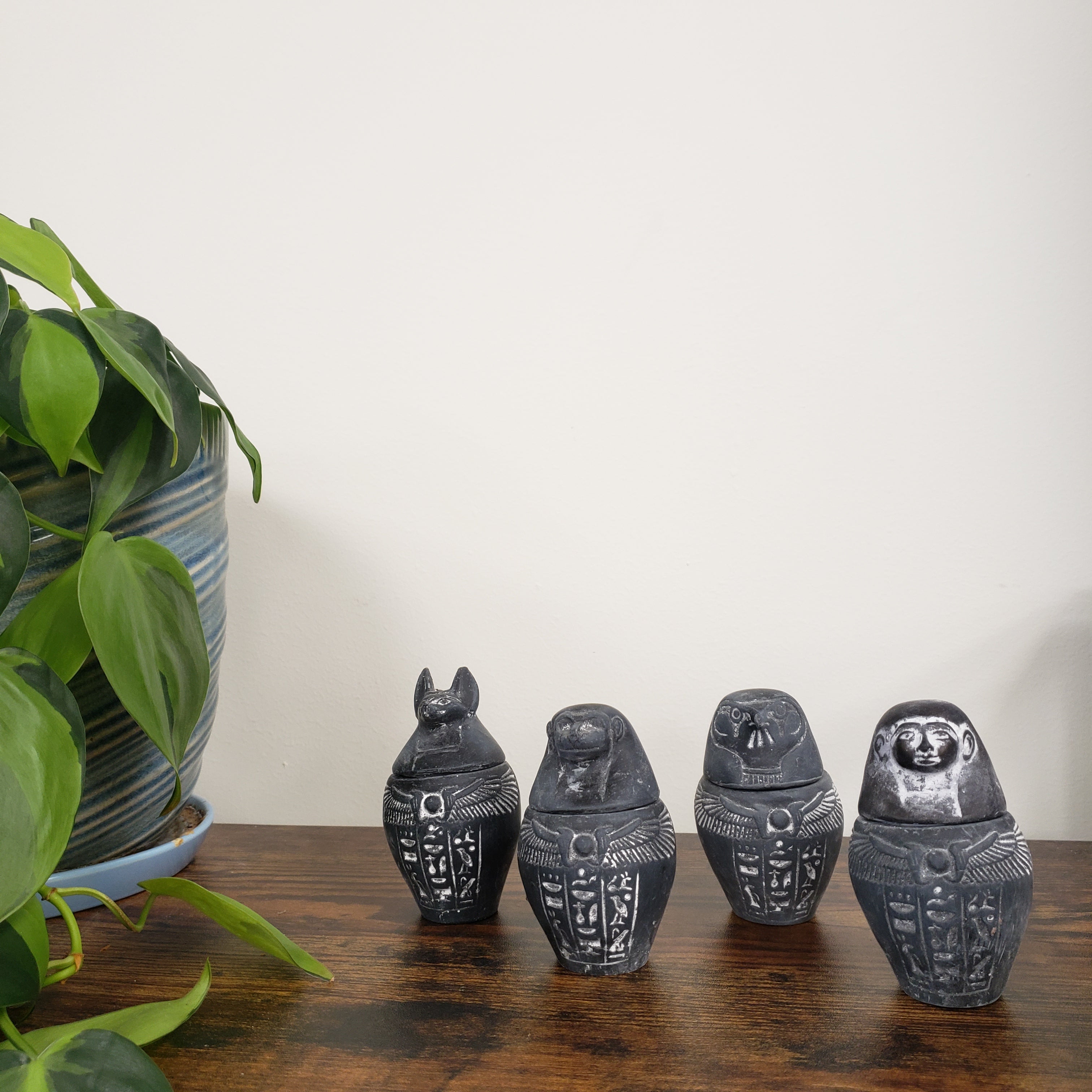 Decorative Egyptian-style canopic jars with intricate designs on a wooden surface with plants in the foreground.