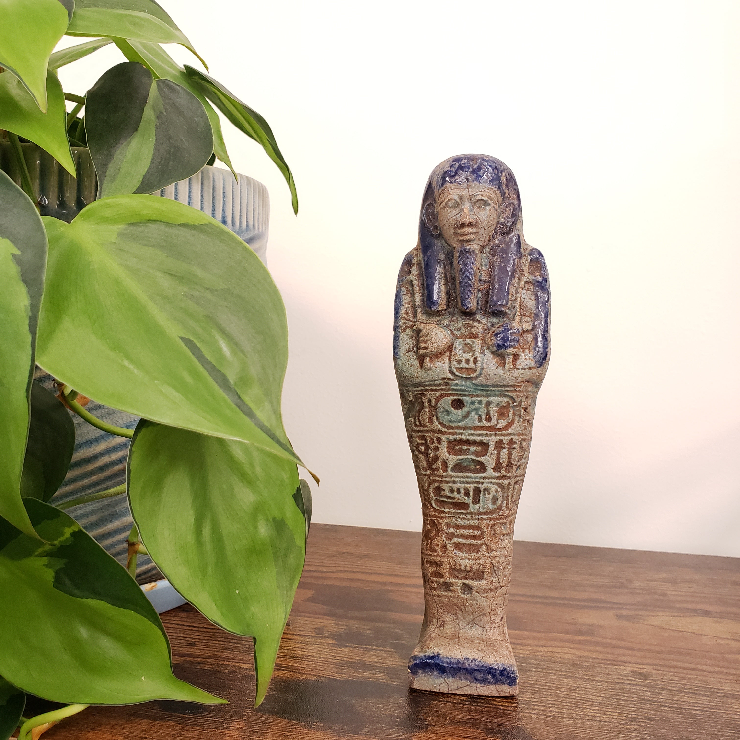 Decorative Egyptian-style ushabti statue with hieroglyphics on a wooden surface with green leaves in the foreground.