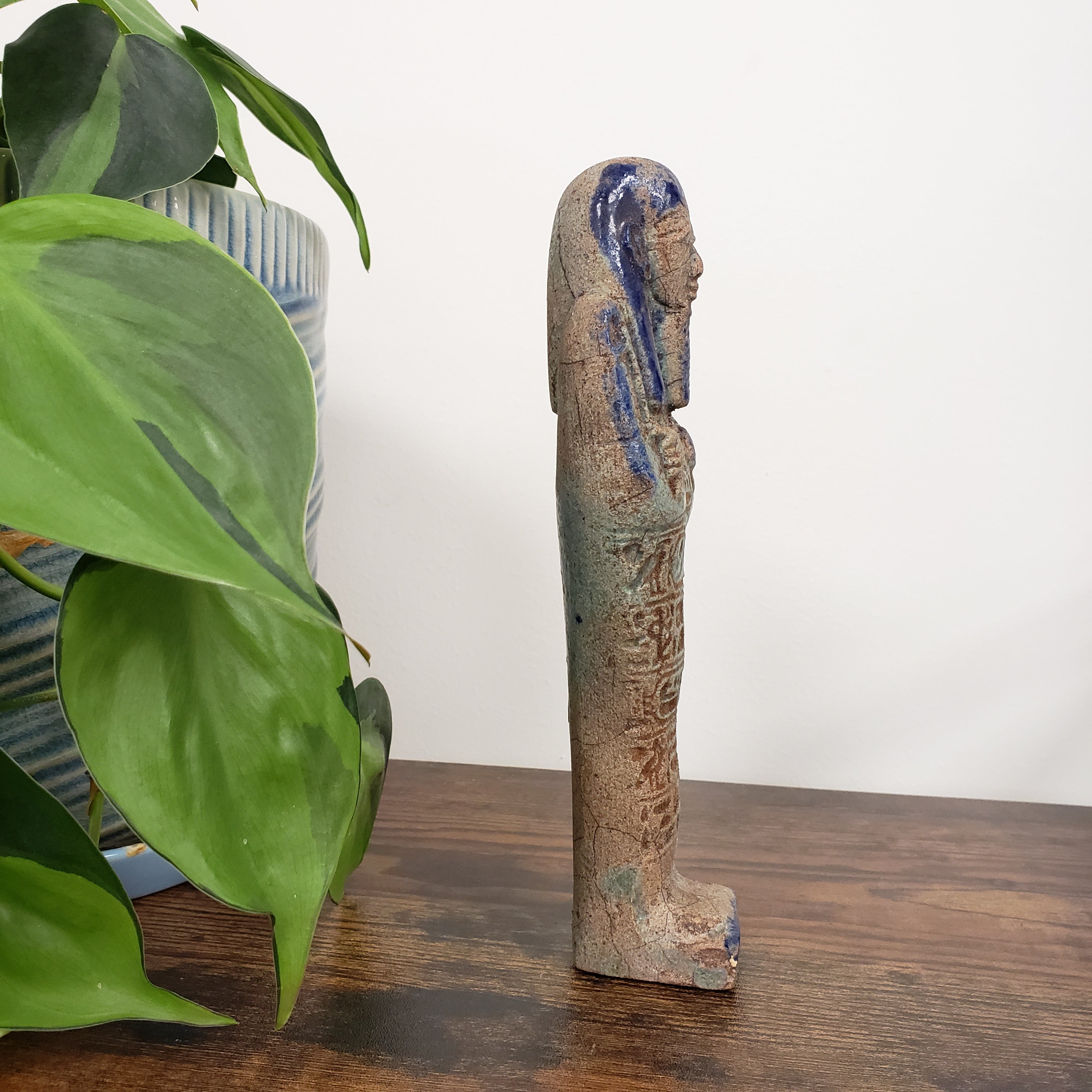 Decorative ushabti statue with blue accents on a wooden surface with green leaves in the foreground.
