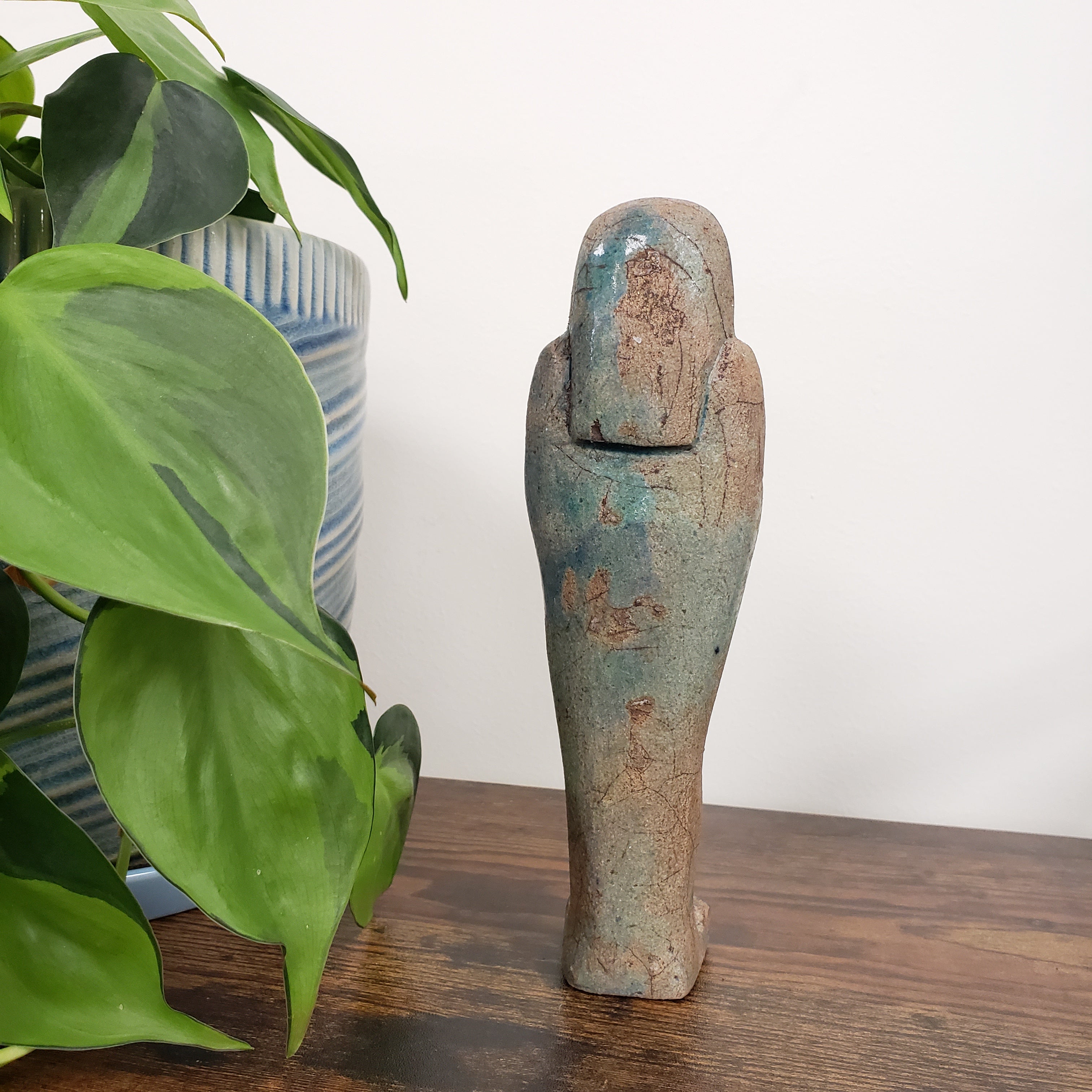 ushabti statue with green patina on a white wall with plants in the foreground