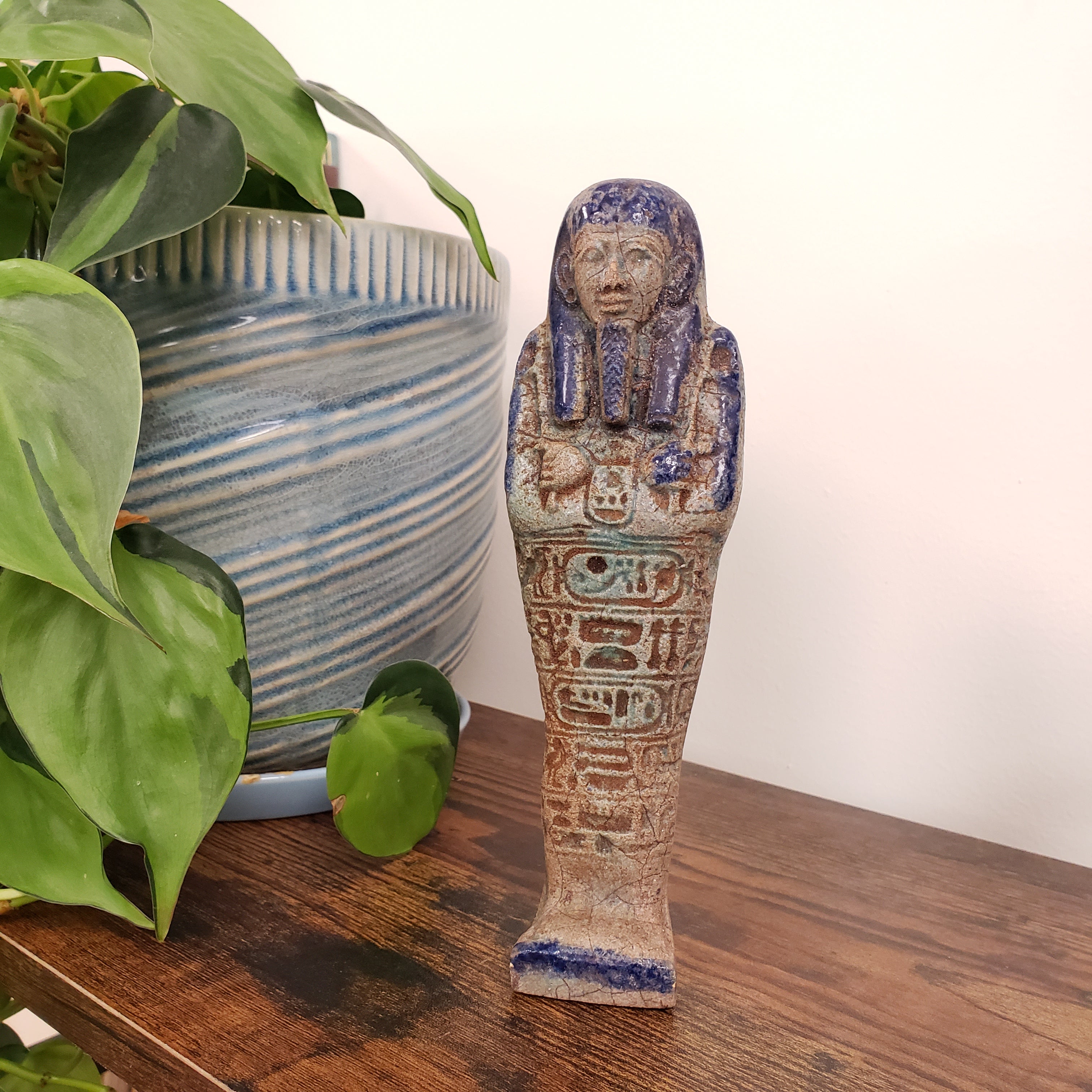 Decorative ceramic ushabti statue with hieroglyphics on a wooden surface with plants in the foreground
