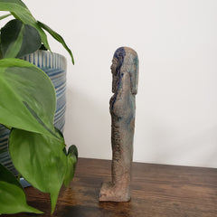 Decorative stone ushabti statue on a wooden surface with green leaves in the foreground
