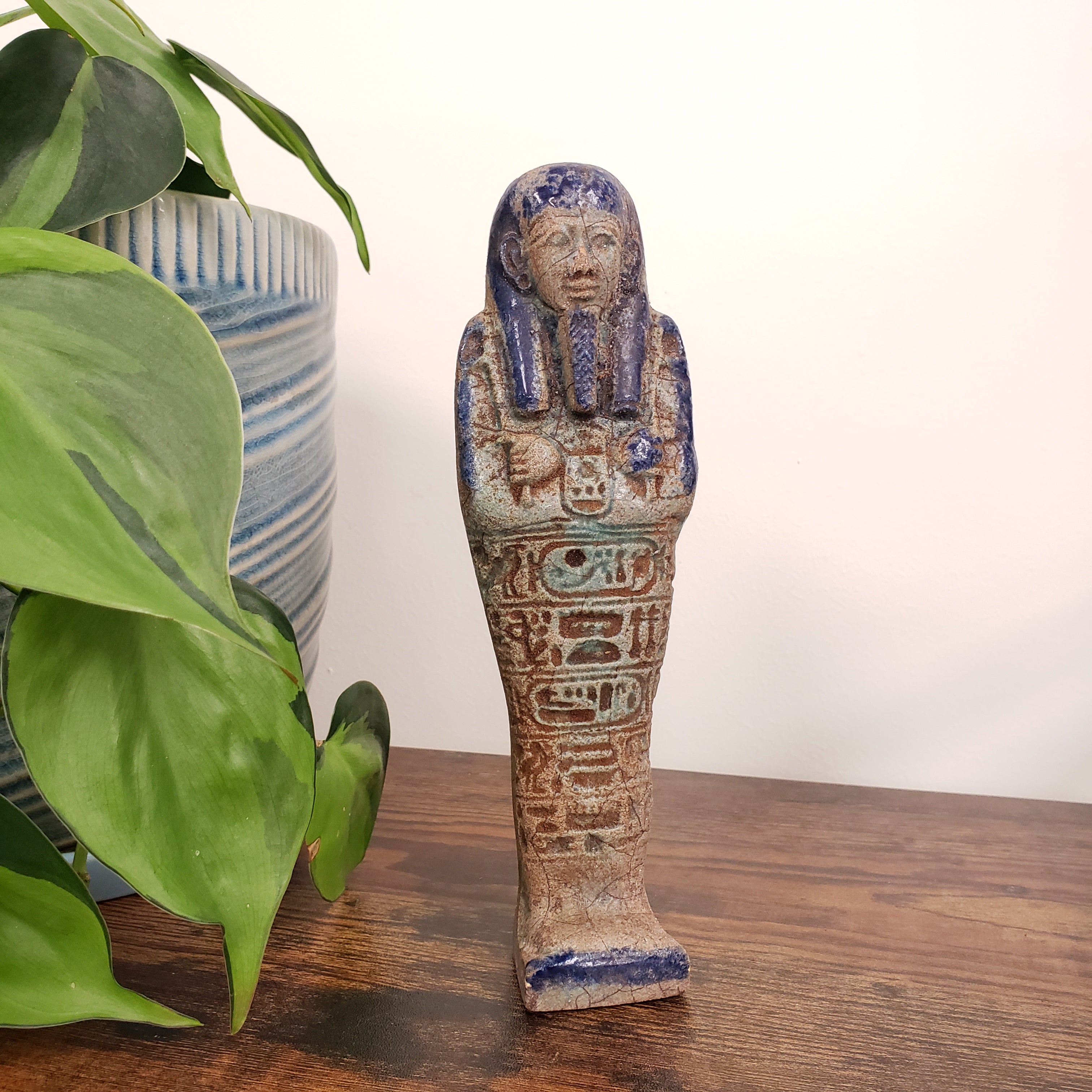 Decorative ceramic ushabti with intricate designs mounted on a wooden wall next to a plant.