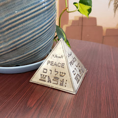 A brass Egyptian pyramid figurine with 'PEACE' written in different scripts, placed on a wooden surface next to a blue vase with a green leaf.
