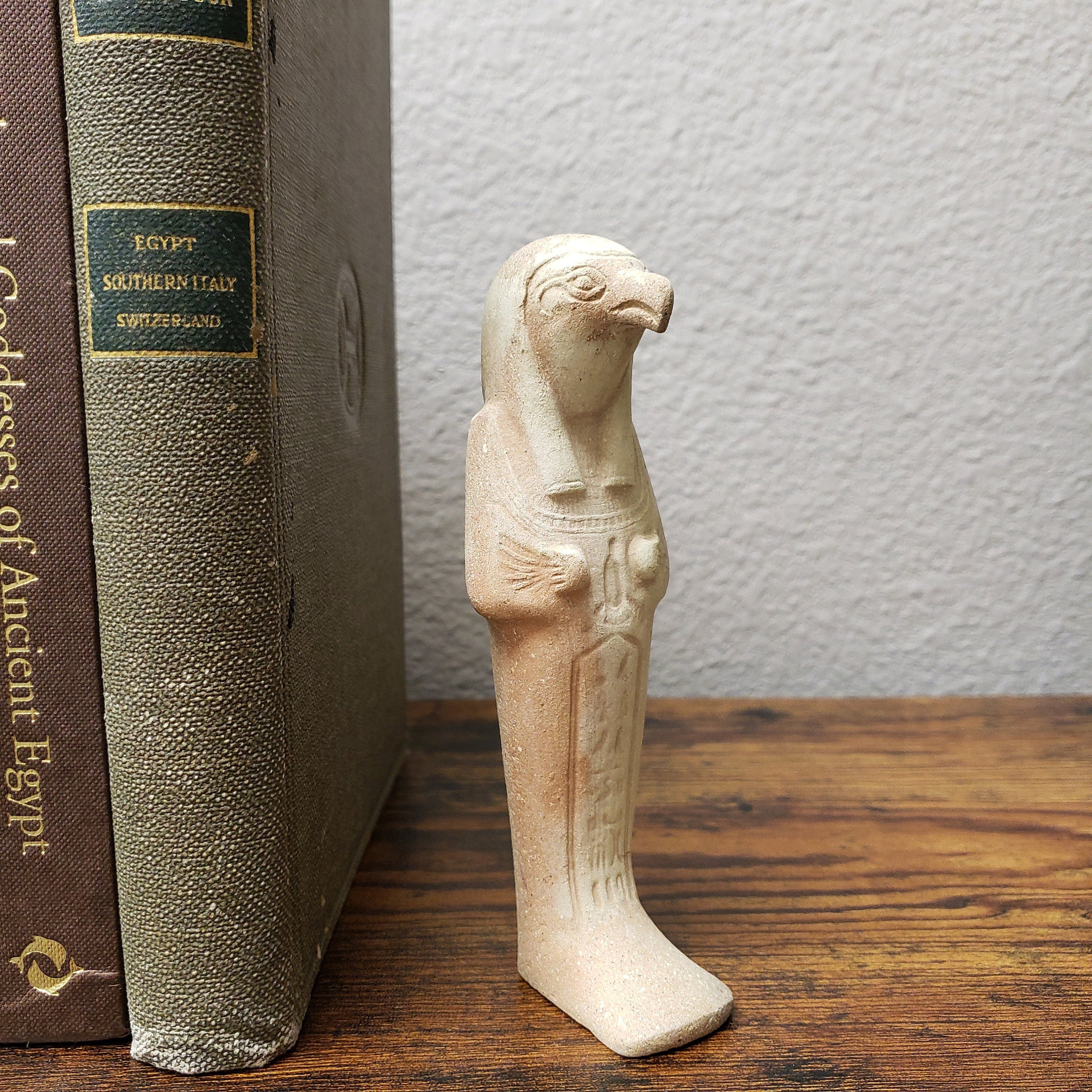 A beige ceramic statue of the Egyptian god Horus, standing next to a book on a shelf.