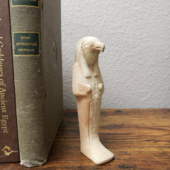 A beige ceramic statue of the Egyptian god Horus, standing next to a book on a shelf.