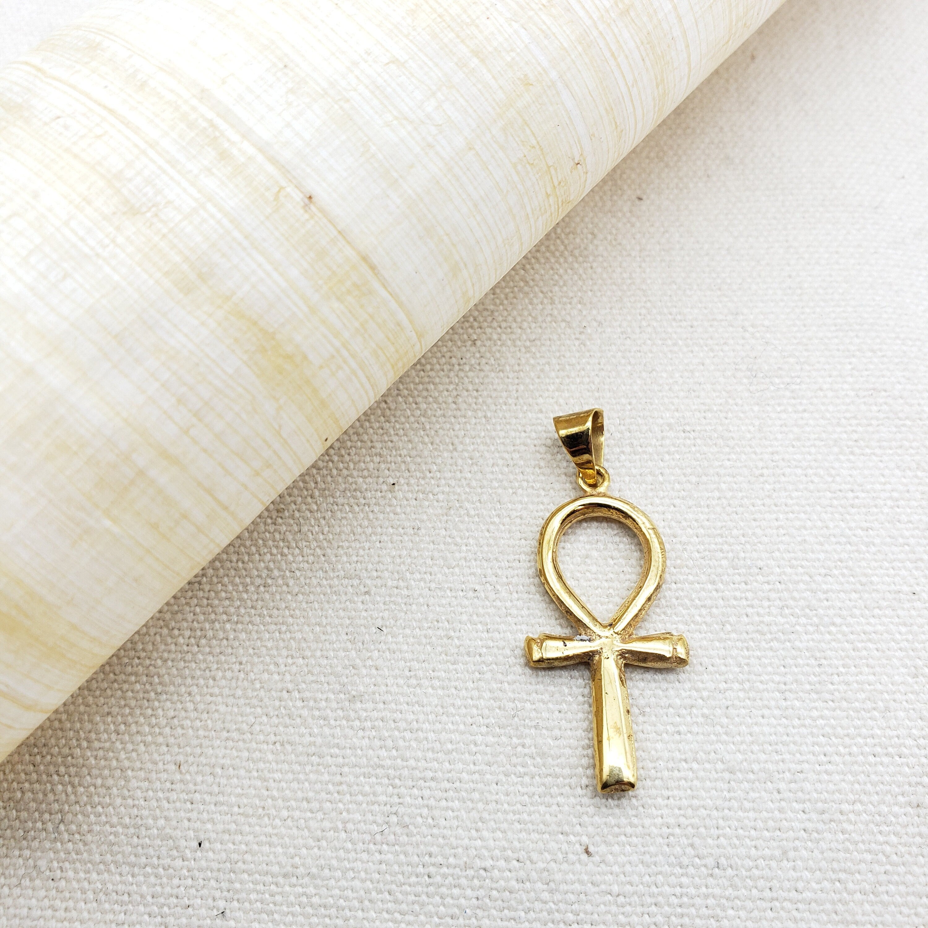 Gold plated ankh pendant next to a white roll of paper on a beige background.